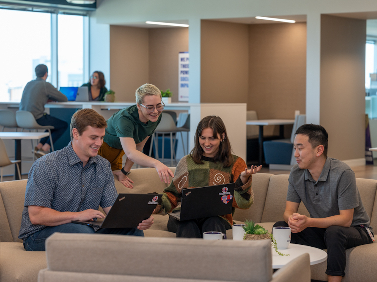 Four Bonterra employees socializing and working. Three are on a corner couch, one is pointing over another person's shoulder while standing behind the couch.