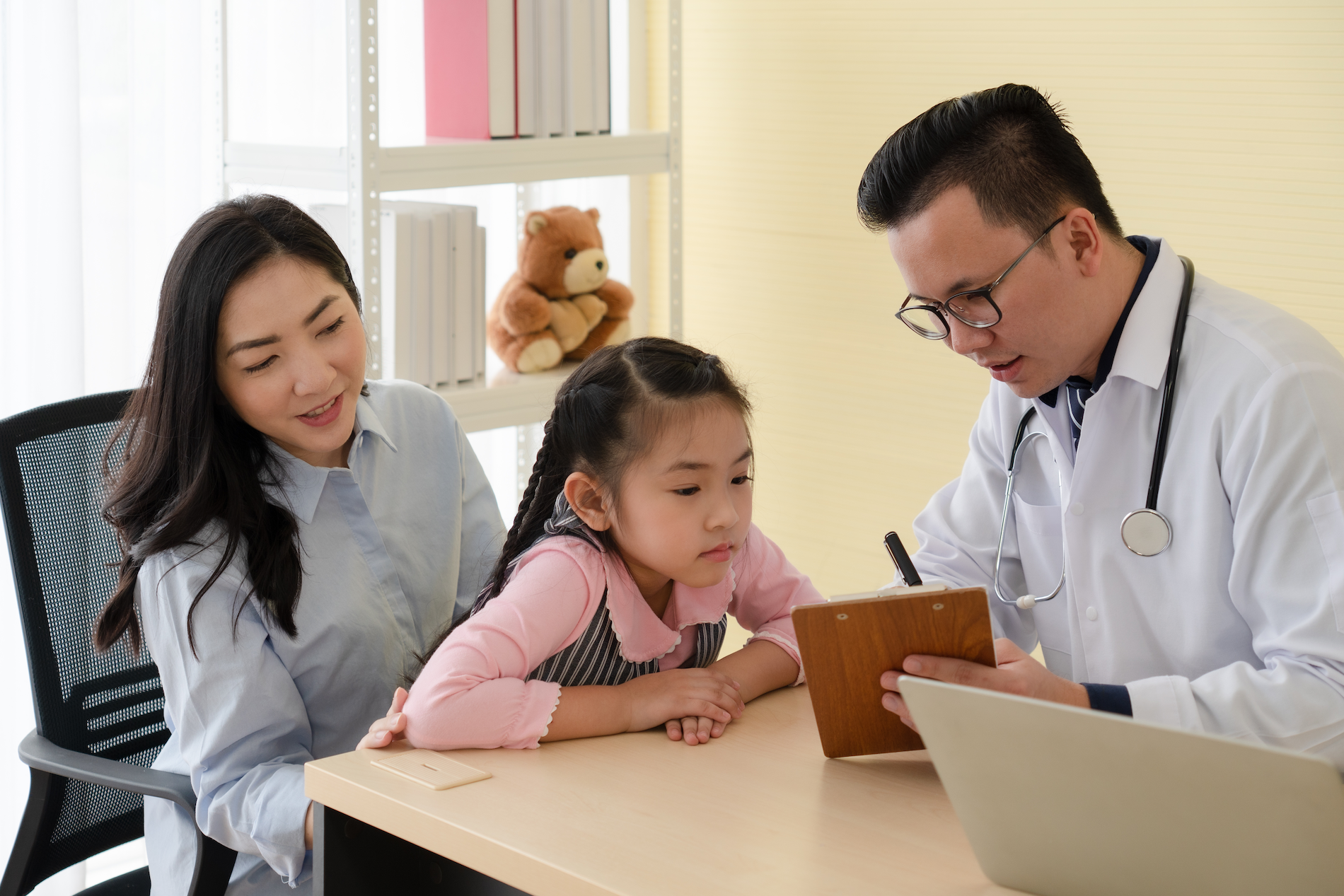 An asian woman sits in a chair with her daughter on her lap. She wears braided pigtails and a pink top. The little girl leans over a desk. An male Asian doctor is showing her something on a notepad.