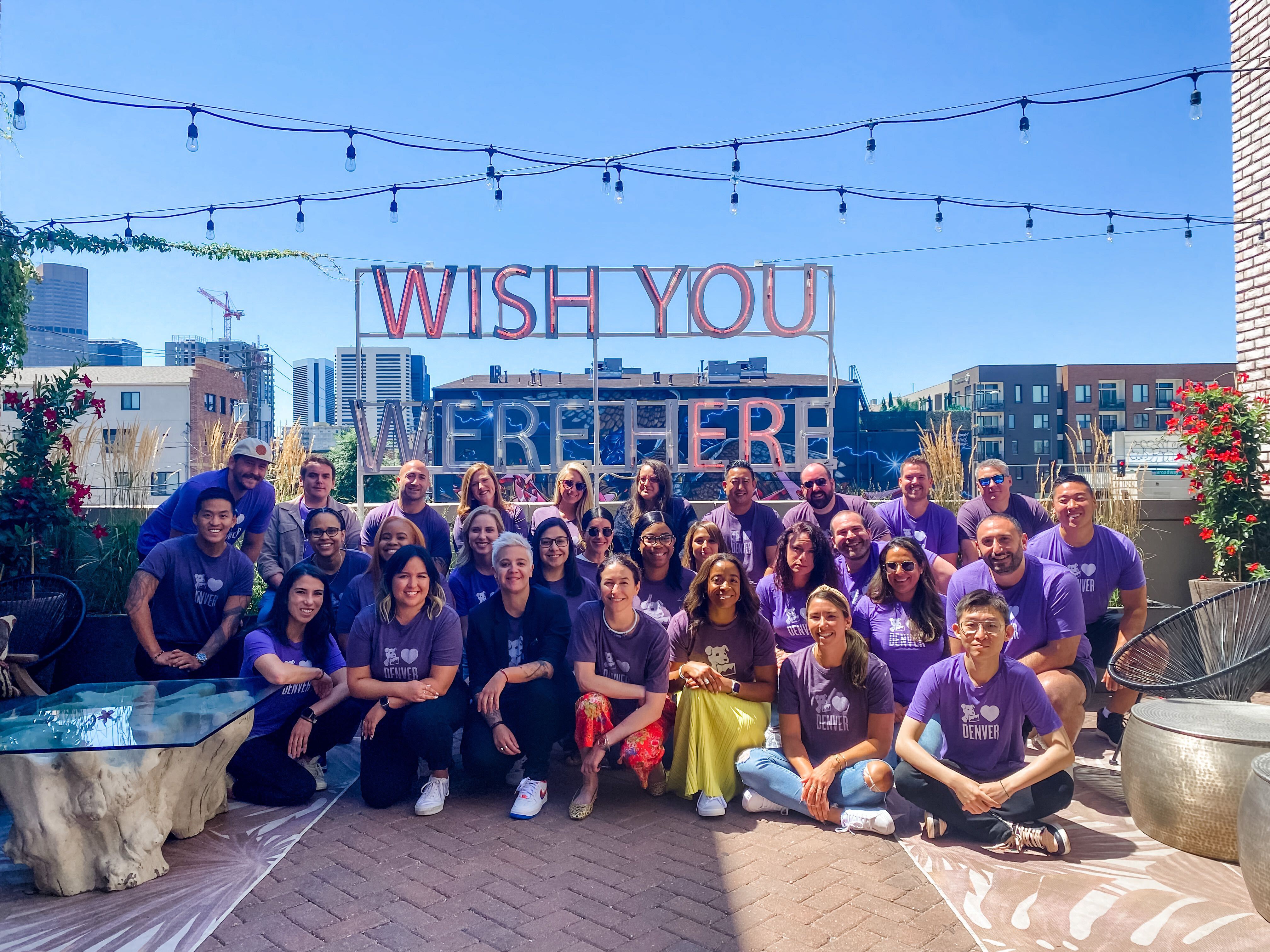 Datadog’s team members sit for a group photo on a rooftop deck with a sign behind them that reads “Wish you were here.”