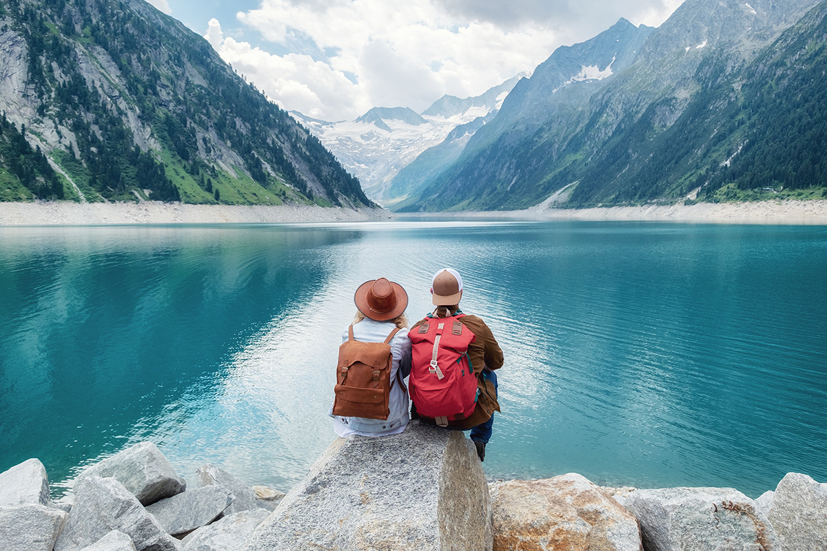 A couple wearing backpacks sitting a rock looking at a lake and mountains