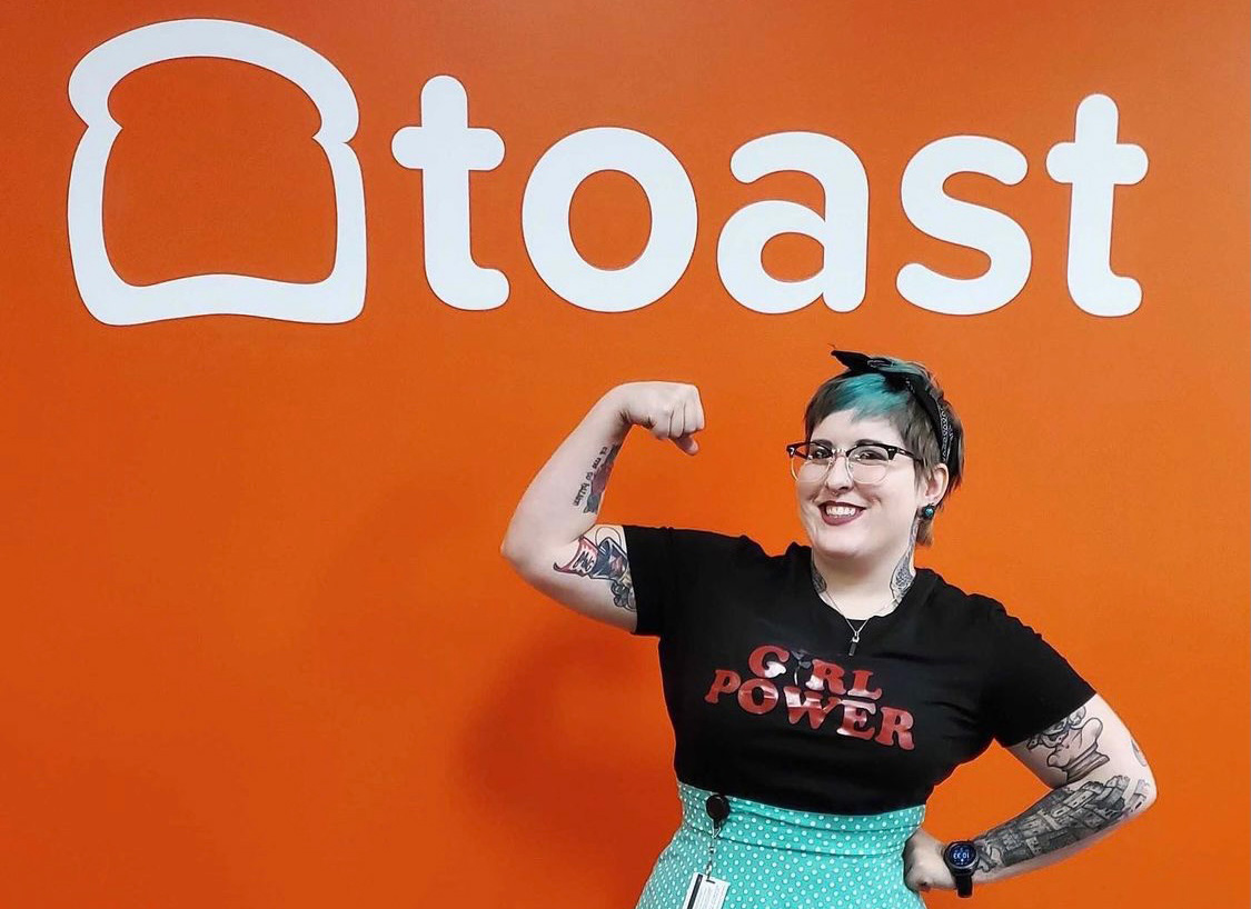 Toast team member wearing a girl power t-shirt flexing her arm muscle in front of an orange wall with the company logo on it
