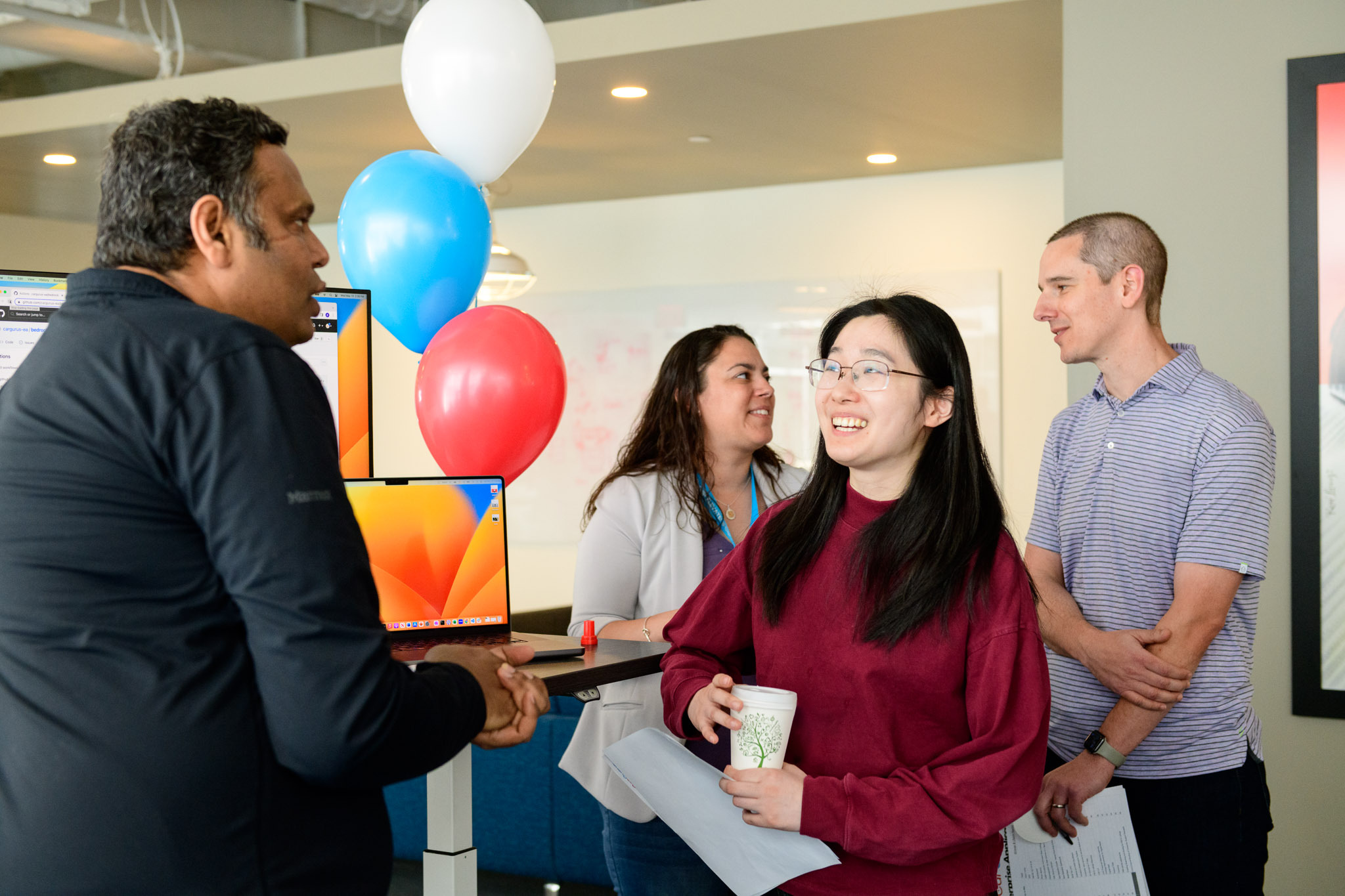 Candid photo of man talking to smiling woman next to computer screens with balloons and colleagues behind her.