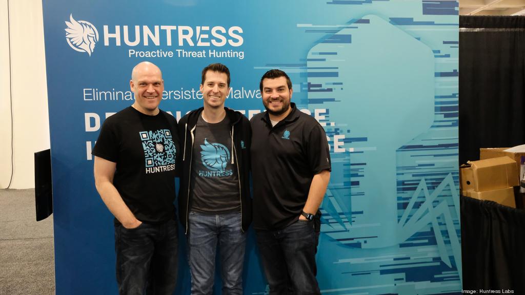 The three Huntress co-founders stand in front of a Huntress sign.