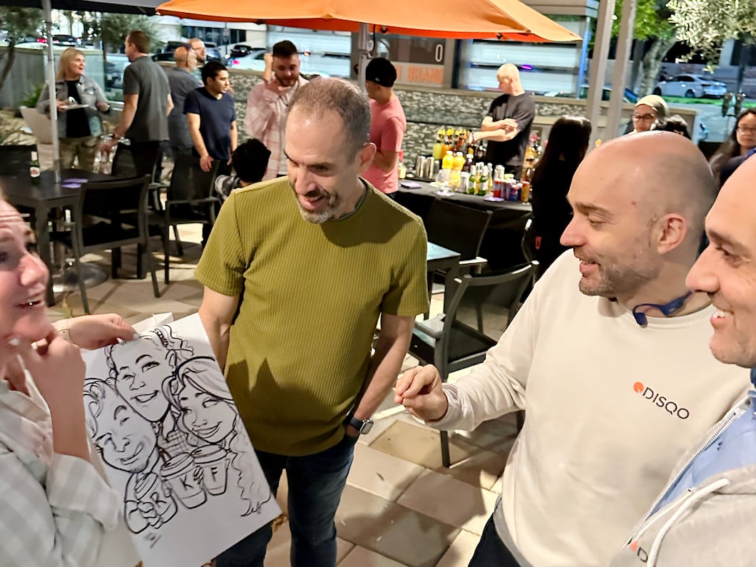 Four DISQO team members at an outdoor party, looking at caricature drawing and smiling, talking.