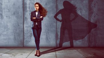 A businesswoman casting a shadow wearing a superhero cape onto the cement wall behind her