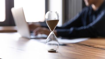 a close up of an hourglass on a wooden table with a person working on a laptop in the background