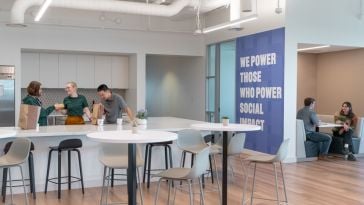 Employees in Bonterra's office. On the right there are two employees at a table. On the left there are three in the kitchen. Between is a wall that reads "WE POWER THOSE WHO POWER SOCIAL IMPACT."
