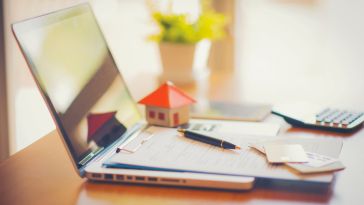 A laptop on a desk with paperwork, credit cards and a house made from paper