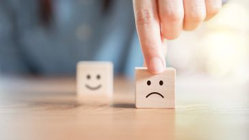 Two wooden blocks, one with a happy face and one with a frown. A human hand is pushing the frown block forward.