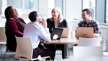 Photo of four colleagues sitting at a table with laptops open, laughing at a joke.