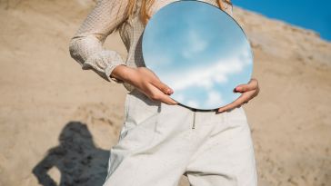 A woman in beige holds a mirror, reflecting the sky.
