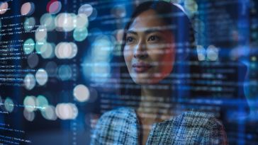 An Asian female stands looking confident and inspired as she studies lines of code on a screen before her eyes