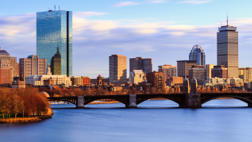 A landscape view of the Boston skyline and harbor. The sunset reflects off the buildings to the west.