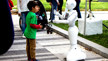 A little boy in a green shirt stands in front of a white robot. They appear to be communicating to one another.